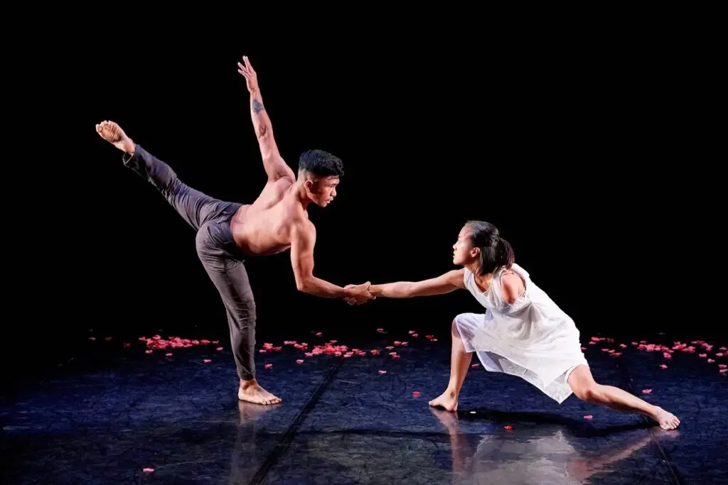 Photo of a choreographed performance featuring a performer on the left reaching out for the hand of another performer.