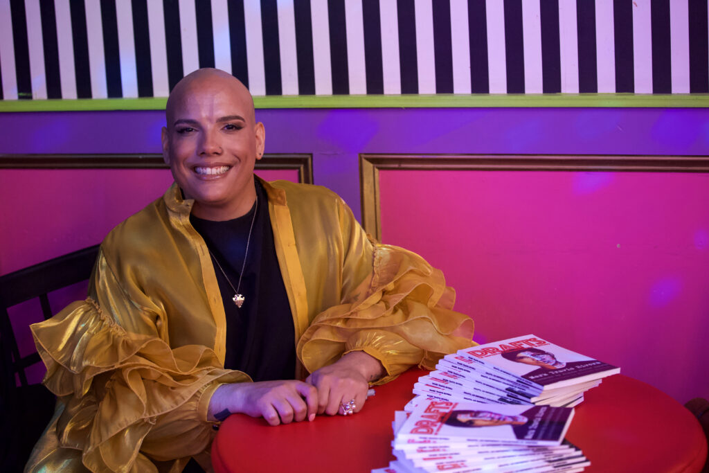 Photo of Mark Travis Rivera smiling whilst sitting at a table, presenting his book, Drafts.