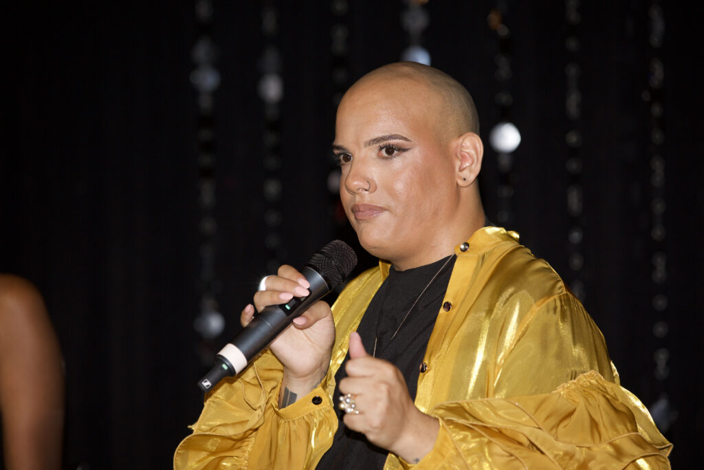 Mark Travis Rivera on stage with a microphone in his hand speaking at Creative Mornings. Mark is wearing a silky yellow top with flared sleeves and a black undershirt.