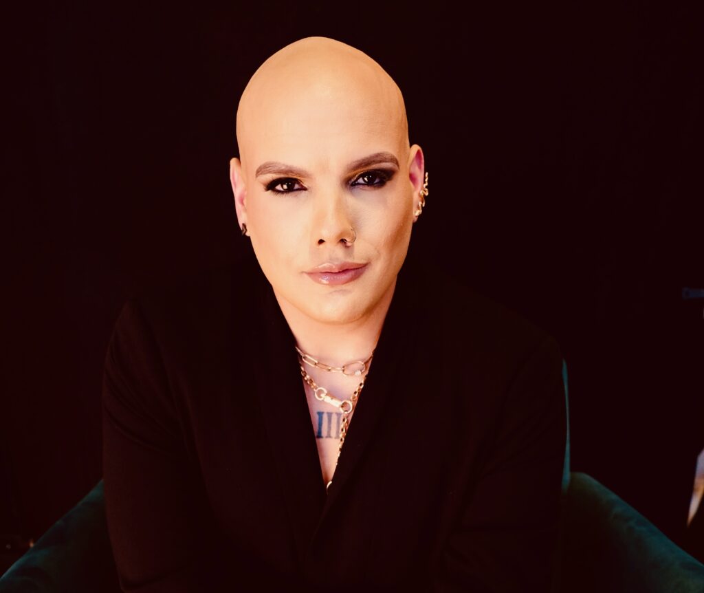 Mark Travis Rivera wearing a black shirt and gold accessories in front of a black back-drop.