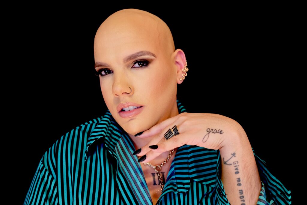 Mark Travis Rivera is looking towards the camera with his hand on his chin. Mark is wearing gold nose and ear accessories, and a blue dress shirt with black vertical stripes in front of a black back-drop.