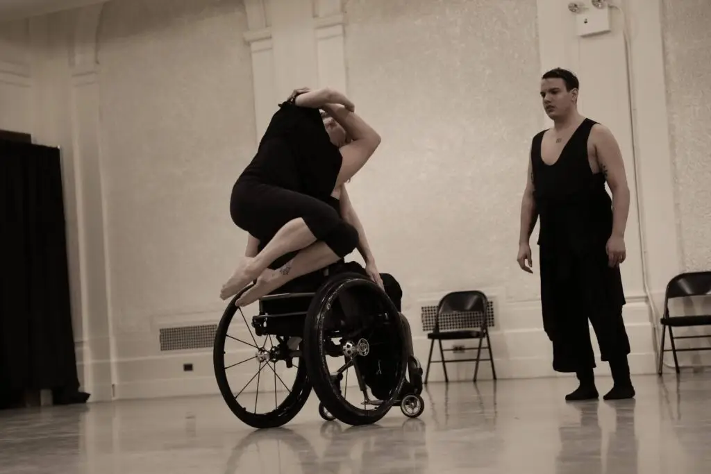 Photo of Mark Travis Rivera watching two dancers practice choreography. One dancer is in a wheelchair holding up another dancer that is leaning on them with their feet bent and tucked and arms crossed above their head.